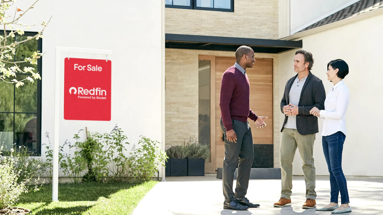 A Redfin agent talking with two homeowners outside a home with a Redfin For Sale sign in the front yard