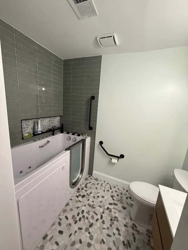 Walk-in jetted tub with sage green tile surround, pebble floor, and grab bars after accessible bathroom remodel in Severn MD